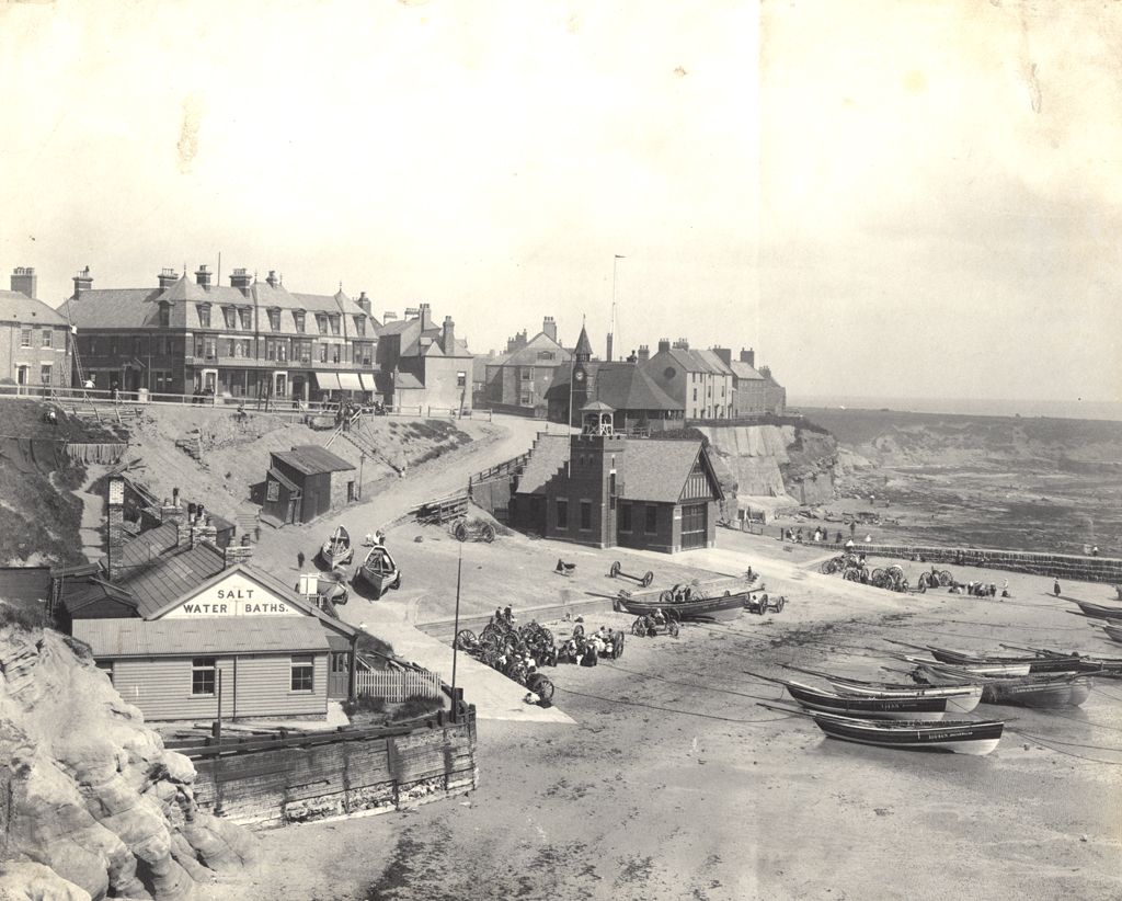 A black and white photo of Cullercaots bay with boats on the beach and a building in the foreground with salt water baths written on it. The lifeboat station can be seen on the opposite side of the bay.