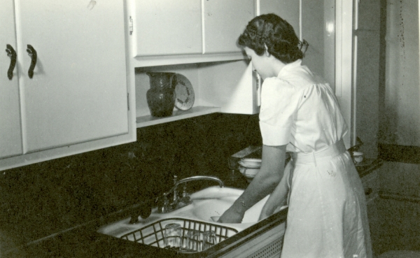 Black and white photo of a young woman at a sink in a kitchen
