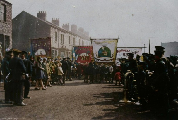 A group of people are walking down a street carrying church banners while others stand at the side of the street watching