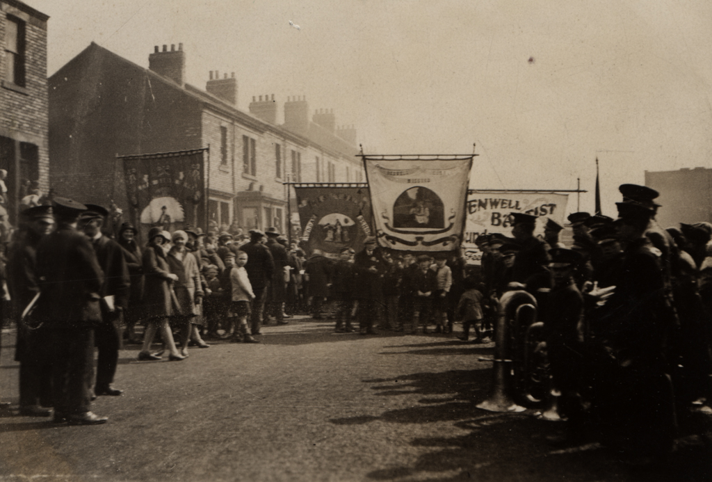 A group of people are walking down a street carrying church banners while others stand at the side of the street watching