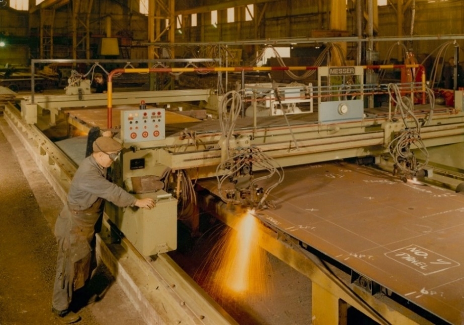 colour photograph of work in the shipyards a worker wearing overalls, a flat cap and dark goggles is operating a machine that is cutting sheets of metal 