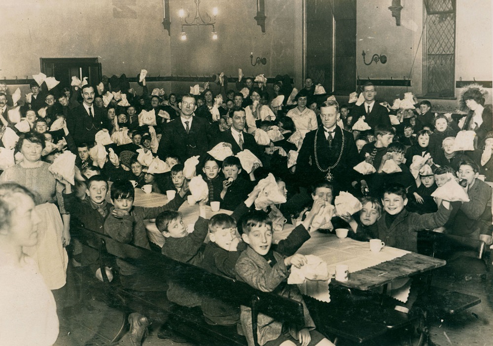 Black and white photo of a large number of children sitting at long tables with adults standing around them. The children are waving bags in the air presumably showing the gifts they have been given at the party